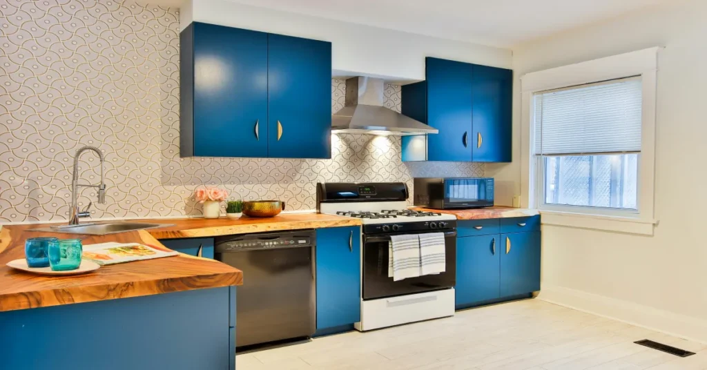 A bright apartment kitchen with vibrant blue cabinets, a live-edge wooden countertop, a stainless steel sink, and a compact dishwasher installed next to a gas stove.