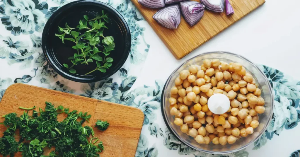 Top-down view of a clear mini food processor bowl filled with chickpeas on a kitchen counter next to chopped fresh herbs and red onions