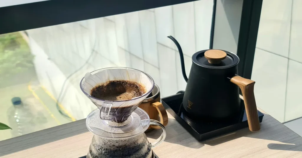 Best Small Electric Kettles for Tight Counter Spaces Matte black electric gooseneck kettle with wooden handle on heating base alongside clear glass pour-over coffee dripper on countertop.